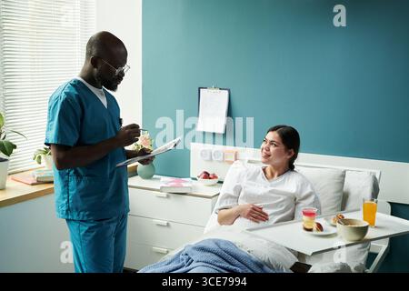 Glückliche schwangere Frau, die im Krankenhausbett liegt und mit der männlichen Krankenschwester spricht Stockfoto
