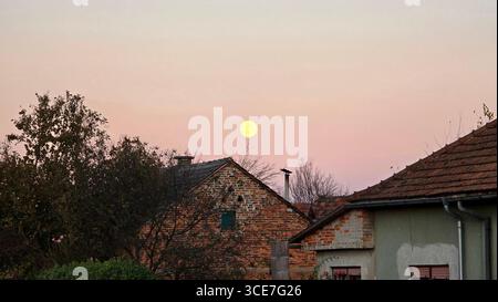 Runder Vollmond erscheint über den Dächern klassischer Bauernhäuser, eingerahmt von blattlosen Herbstzweigen und glühend in sanftem pastellfarbenem Abendhimmel Stockfoto