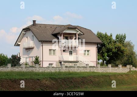 Helles zweistöckiges Haus mit mehreren weißen Balkonen, geneigtem Dach und Steinmauer, umgeben von üppigen Gartenbäumen auf einer klaren Sonne Stockfoto
