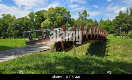 Sanft geschwungener Holzsteg mit Stahlgittergeländern verbindet Wanderwege über einen flachen, grasbewachsenen Graben, umgeben von alten Bäumen in sonniger Stadt Stockfoto