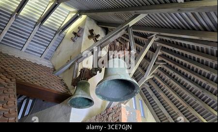 Zwei alte Bronzeglocken hängen in einem überdachten hölzernen Glockenturm, umgeben von freiliegenden Balken, Mauerwerk und traditionellen Lehmbachziegeln Stockfoto