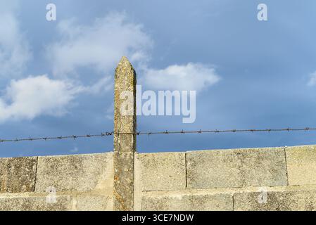 Betonmauer mit Stacheldraht gegen einen bewölkten Himmel Eine Sicherheitsbarriere aus Betonblöcken und scharfem Draht unter einem dramatischen Himmel. Stockfoto