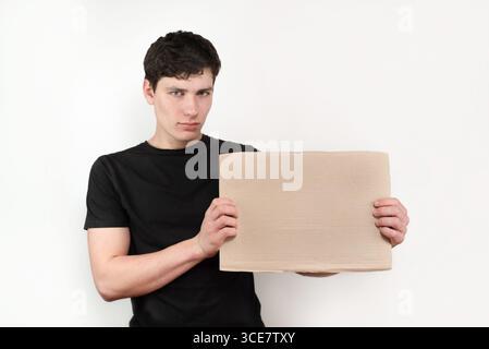 Junger Erwachsener, der ein schwarzes Hemd trägt und ein leeres braunes Pappschild hält, perfekt für individuelle Anpassungen. Vor weißem Hintergrund aufgenommen Stockfoto