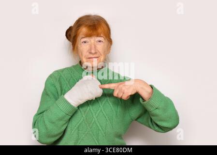 Schlanke Frau in einem grünen Pullover mit roten Haaren, die auf ihre bandagierte Hand vor einem glatten, weißen Hintergrund zeigen. Die Frau stolperte und fiel Stockfoto