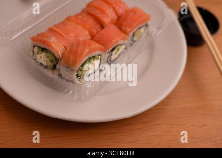 Frisch zubereitete Sushi-Lachsbrötchen auf einem weißen Teller, begleitet von Essstäbchen und einer Seite Dippsoße auf einem Holztisch Hintergrund Stockfoto