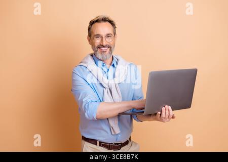 Ein selbstbewusster, reifer Geschäftsmann, der ein Notebook benutzt, während er vor einem beigefarbenen Hintergrund steht und professionelle Eleganz zeigt Stockfoto