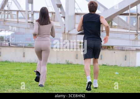 Junger Mann und Frau, die zusammen im Grünen Gras unter einer großen Brücke joggen Stockfoto