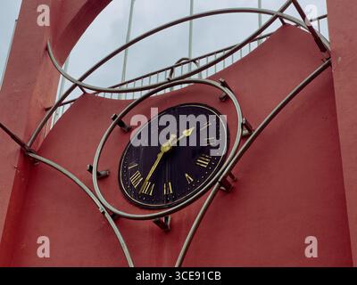 Detaillierte Ansicht einer großen Uhr in Schwarz und Gold mit römischen Ziffern, die in eine markante rote Wand eingefasst und von modernen Metallstrukturen eingerahmt ist. Stockfoto
