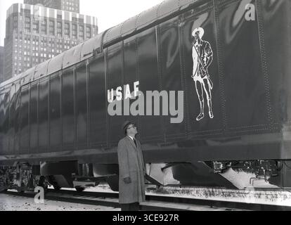 1950er Jahre, historisch, draußen auf schneebedecktem Boden an einer Bahnstrecke, ein Gentleman mit Mantel, Handschuhen und Hut, der auf einen großen Eisenbahnwagen blickt, mit den Buchstaben U.S.A.F. an der Seite und einer Figur eines Soldaten aus dem 18. Jahrhundert, Chicago, USA. Stockfoto