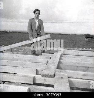 1950er Jahre, historisch, ein neuer Bungalow im Bau, Bild zeigt einen Handwerker, einen jungen Mann, der auf den Holzbalken eines neuen Daches steht und einen Teil des Holzes hält. Stockfoto