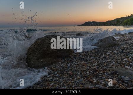 Ein ruhiger Blick auf die Wellen, die gegen zerklüftete Felsen plätschern und die ruhige Essenz eines Sonnenuntergangs am Strand einfangen, mit sanften Farben, die sich im Himmel A verschmelzen Stockfoto