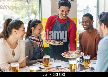 Freunde genießen frische Pizza und Bier in einem gemütlichen Restaurant. Konzept der Freundschaft, multikultureller Zusammenkunft, zwangloses Essen und authentisches italienisches Essen e Stockfoto