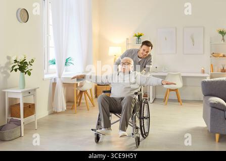 Junge freundliche Krankenschwester schiebt glücklichen älteren behinderten Mann im Rollstuhl, der Spaß im Pflegeheim hat. Stockfoto