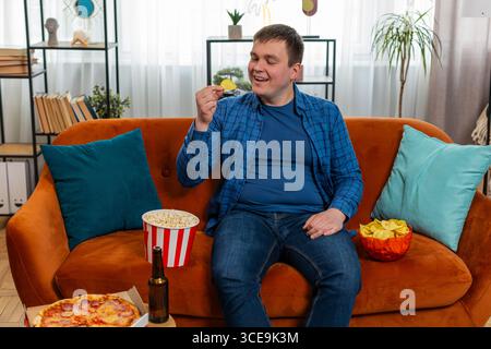 Glücklicher junger Mann in ungezwungener Kleidung, der auf einem bequemen Sofa sitzt und Chips, Pizza, Popcorn und Bier trinkt, um Filmsport zu beobachten und ein Wochenende zu Hause zu verbringen. Ein lächelnder Kerl isst Junk Food auf der Couch im Haus. Stockfoto