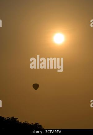 Heißluftballon, der bei Sonnenaufgang unter der Sonne vorbeizieht, Silhouette gegen den Himmel, aufgenommen auf den Somerset Levels, Großbritannien Stockfoto