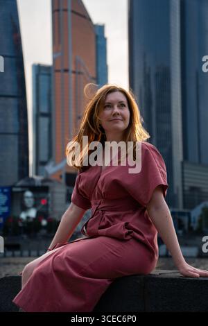 Frau in rotem Kleid sitzt am Fluss mit Wolkenkratzern von Moskau-Stadt im Hintergrund, sanft beleuchtet durch Sonnenuntergang Stockfoto