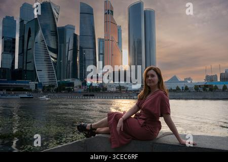 Frau in rotem Kleid sitzt am Fluss bei Sonnenuntergang mit Wolkenkratzern von Moskau im Hintergrund Stockfoto