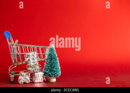 Kleiner Einkaufswagen mit glänzenden silbernen Geschenkboxen vorne und grünem Weihnachtsbaum auf roter Oberfläche mit tiefrotem Hintergrund. Konzept von fe Stockfoto