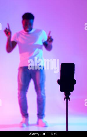 Schwarzer männlicher Influencer, der Video für soziale Medien auf dem Mobiltelefon dreht, Spaß im Neonlicht hat, Nachbau Stockfoto