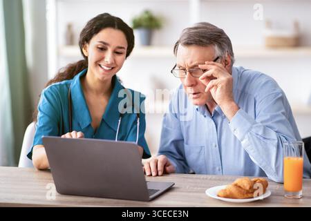 Junge Krankenschwester zeigt älteren behinderten Mann, wie man Laptop zu verwenden Computer im Altersheim Stockfoto