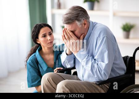 Depressiver älterer Mann im Rollstuhl weinend, junger Arzt versucht, ihm im Altersheim zu helfen Stockfoto