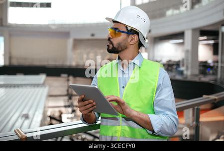 Serious nachdenklich tausendjährigen islamischen männlichen Ingenieur in Schutzuniform, Brille und Hut mit Bart mit Tablette Stockfoto