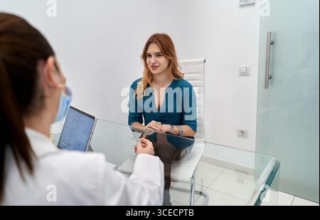 Ein freundlicher Zahnarzt in weißem Mantel und Maske berät eine lächelnde Patientin in einer modernen Zahnklinik. Die Umgebung ist sauber und einladend Stockfoto