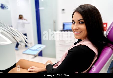 Eine Frau sitzt bequem in einem Zahnarztstuhl und lächelt selbstbewusst in die Kamera in einer modernen Zahnklinik. Der Hintergrund zeigt eine hygienische und professionelle Stockfoto
