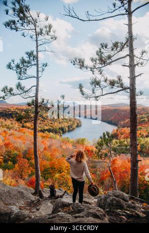 Eine Frau steht auf einem Hügel mit Blick auf eine atemberaubende Herbstlandschaft in Quebec, Kanada. Das lebhafte Herbstlaub und der ruhige See unter Ihnen sorgen für einen Atem Stockfoto