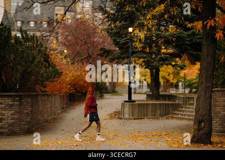 Seitenansicht einer nicht erkennbaren rothaarigen Frau, die durch eine lebhafte Herbstszene in Montreal läuft. Der Weg ist gesäumt von bunten Bäumen und gefallenen Leaves Stockfoto