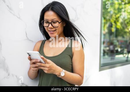 Eine junge Chinesin steht draußen in einer Sommerstadt und lächelt, während sie ihr Smartphone benutzt. Sie ist lässig gekleidet und genießt einen sonnigen Tag in t Stockfoto