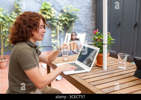 Seitenansicht eines jungen Mannes, der an einem Laptop arbeitet, in einem lebhaften Außenbereich, umgeben von Pflanzen und natürlichem Licht, schafft eine friedliche und professionelle Atmosphäre Stockfoto