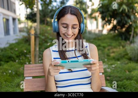 Eine junge Frau entspannt sich auf einer Bank im Stadtpark, trägt Kopfhörer und spielt eine Spielkonsole. Sie liebt das urbane Outdoor-Leben und genießt Ruhe Stockfoto
