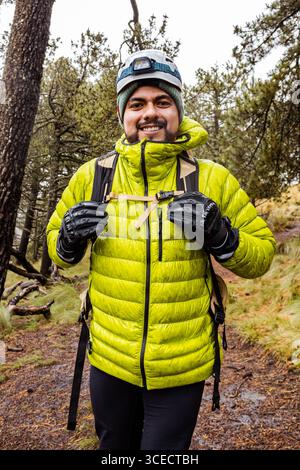 Ein Wanderer in leuchtend grüner Kleidung navigiert auf einem matschigen, regendurchfluteten Pfad durch einen dichten Wald. Ausgerüstet für Abenteuer, nimmt der Wanderer die Herausforderung an Stockfoto