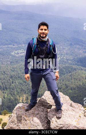 Ein Wanderer steht selbstbewusst auf einer felsigen Klippe, umgeben von einem atemberaubenden bergpanorama. Die mehrschichtige Waldlandschaft und der bewölkte Himmel schaffen Dramati Stockfoto