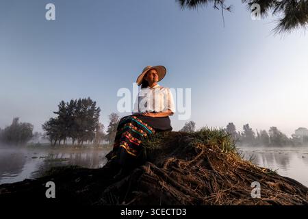 Eine Frau in traditioneller Kleidung genießt einen ruhigen Moment am Wasser in Xochimilco, Mexiko. Umgeben von der Natur spiegelt sie die reiche Kultur der Region wider Stockfoto