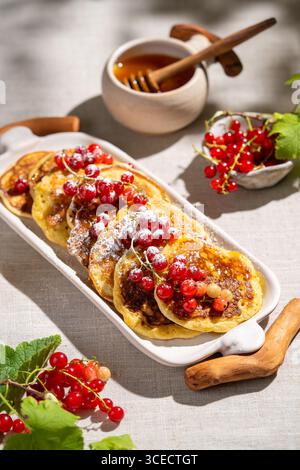 Von oben, eine weiße Platte mit goldenen Pfannkuchen, gekrönt mit leuchtenden roten Johannisbeeren und mit Puderzucker bestäubt auf einem hellen Leinenstoff Hintergrund, Accom Stockfoto