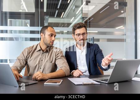 Kollegen arbeiten in einer modernen Büroumgebung zusammen und nutzen Laptops und andere digitale Geräte für Diskussionen. Das Bild hebt Teamwork, Technologie und professionelle Kommunikation hervor Stockfoto