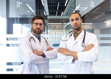 Zwei männliche Mediziner stehen mit überkreuzten Armen, tragen weiße Mäntel und Stethoskope und strahlen Vertrauen und Professionalität in einem modernen Büro aus. Dieses Bild symbolisiert Gesundheit, Teamarbeit und Vertrauen. Stockfoto