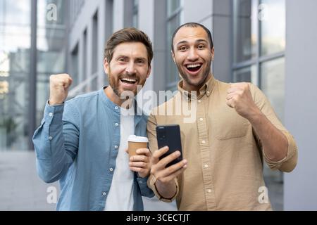 Zwei fröhliche Einzelpersonen stehen in einem Stadtgebiet und feiern gemeinsam mit Enthusiasmus, während sie spannende Nachrichten auf einem Smartphone ansehen. Man hält eine Kaffeetasse, während sie den freudigen Moment teilen. Stockfoto