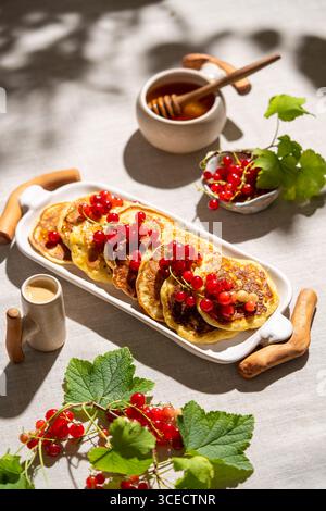 Von oben sind goldene Pfannkuchen mit roten Johannisbeeren wunderschön auf einem weißen Tablett angeordnet. Die umliegenden Gegenstände befinden sich auf einem strukturierten, hellbeigen Leinen Stockfoto