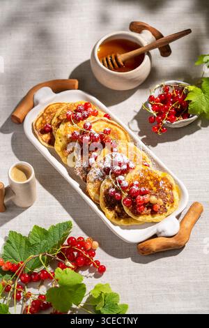 Blick von oben auf köstliche Pfannkuchen mit roten Johannisbeeren auf einem weißen Keramiktablett. Der helle Leinenhintergrund sorgt für eine Frische mit Honig und Cof Stockfoto