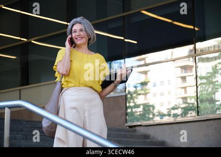Reife Frau in einer gelben Bluse, die am Telefon spricht, während sie vor einem Bürogebäude steht. Sie hält ein Tablet und wirkt selbstbewusst und engagiert Stockfoto