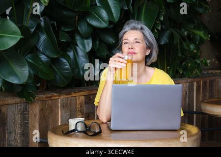 Eine reife Frau sitzt an einem Holztisch mit Laptop und genießt ein erfrischendes orangenes Getränk. Sie strahlt Ruhe und Selbstvertrauen aus, umgeben von üppigem Grün Stockfoto