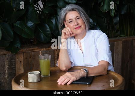 Reife Frau mit kurzen grauen Haaren, die an einem rustikalen Holztisch sitzt und eine nachdenkliche Pose hält. Ein Glas grüner Saft in der Nähe sorgt für eine erfrischende Atmosphäre Stockfoto