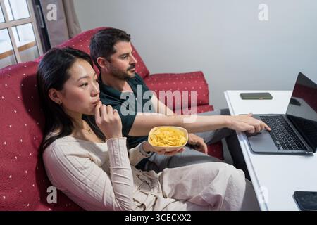 Ein vielseitiges Paar sitzt bequem auf einer Couch zu Hause, nimmt sich mit einem Laptop an und teilt sich eine Schüssel mit Snacks, um ein gemütliches und entspanntes atmos hervorzuheben Stockfoto