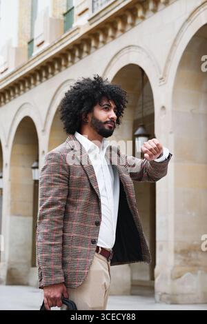Ein Versicherungsverkäufer mit Afro-Haaren, der einen karierten Blazer und ein weißes Hemd trägt, schaut seine Uhr, während er vor einem Steingebäude steht Stockfoto