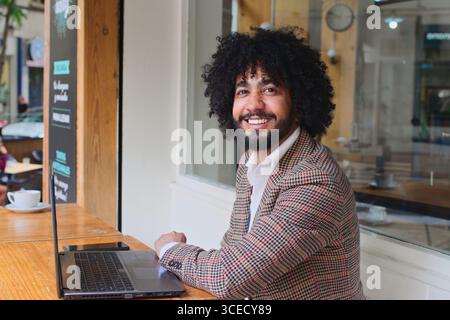 Ein Versicherungsverkäufer mit Afro-Haaren lächelt an einem Holztisch und arbeitet an einem Laptop. Die Inneneinrichtung umfasst ein Café mit warmer Beleuchtung und Stockfoto