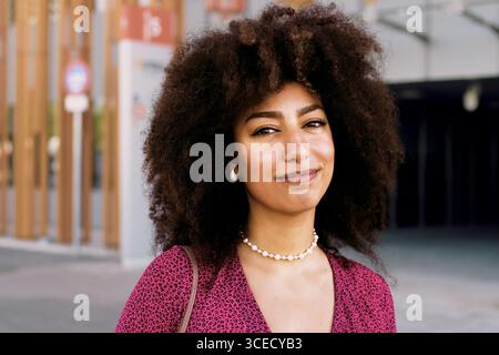 Junge arabische Frau mit wunderschönem Afro-Haar, die selbstbewusst in die Kamera blickt, während sie ein pinkfarbenes Oberteil und eine Perlenkette trägt, die im Outdo steht Stockfoto
