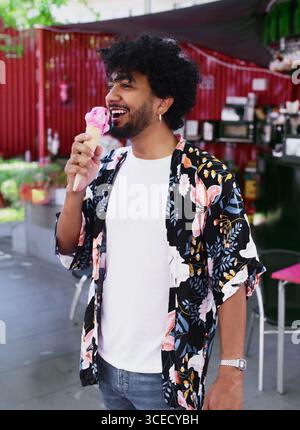 Ein fröhlicher Mann isst ein erfrischendes Eis, während er einen sonnigen Sommertag genießt. In lässiger Kleidung steht er im Freien, umarmt Stockfoto